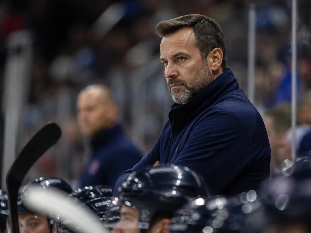 Ishockeytränare i klubbjacka står bakom spelarbänken under en match